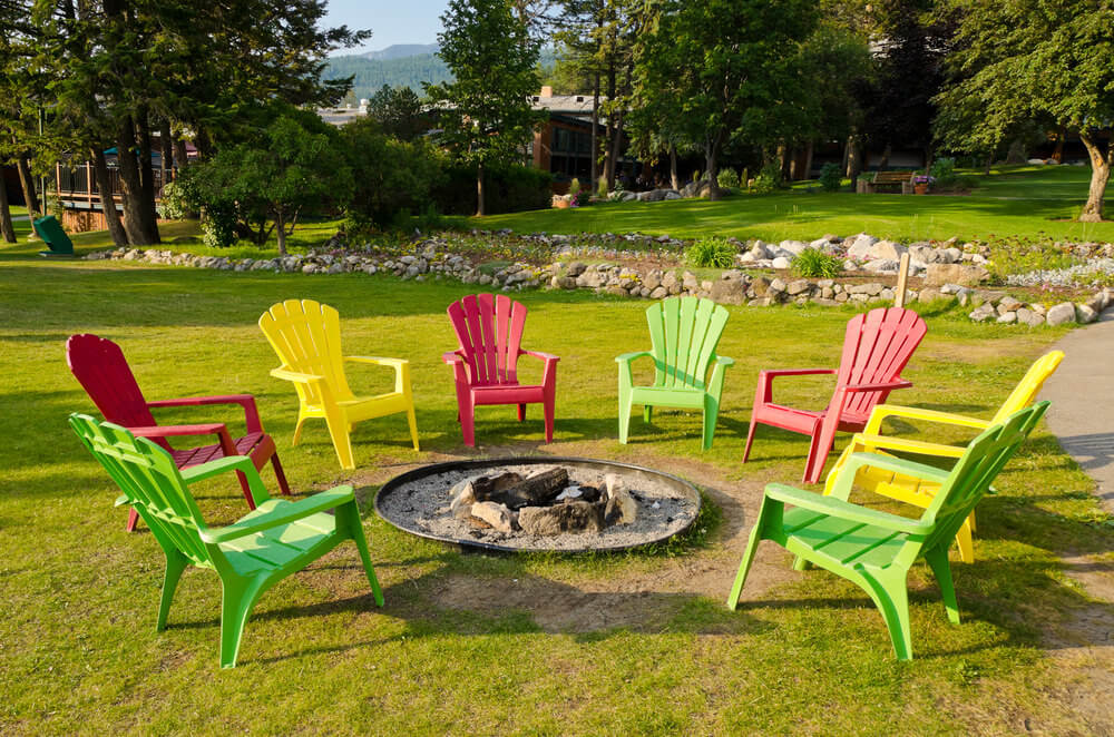 Dug in round fire pit with sand on grassy area surrounded by colourful Adirondack chairs. 在圆形火坑里挖,周围是草地上的沙子,周围是五颜六色的阿迪朗达克椅子。