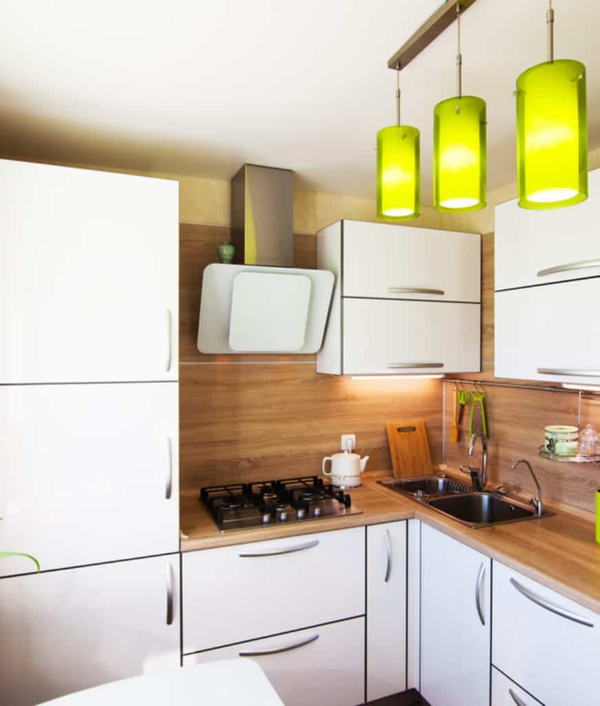 绿色圆柱形吊坠在厨房中脱颖而出，展示了带有铬拉杆的白色橱柜和配有双水槽和灶台的木制柜台。