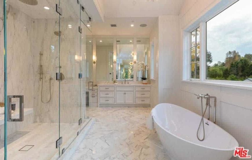 这个浴室空间的设计正吸引着你潜入其中，放松身心。在设计浴室时，使用较浅的颜色总是安全的，因为它确实鼓励了清洁的本质，特别是当你有大窗户融入空间时。我们在地板上有令人惊叹的石头瓷砖，引导您走向现代的蛋形浴缸。一个景观窗可以让你快速一瞥户外的绿色植物，透明的淋浴墙确实提供了一种开放的感觉。我们在后面的墙上有一个古雅而华丽的梳妆台，有古董的破旧橱柜和镜子。这个房间里的一切都是优雅的，经典的，和恰到好处的空间。一个真正独特的浴室设计!