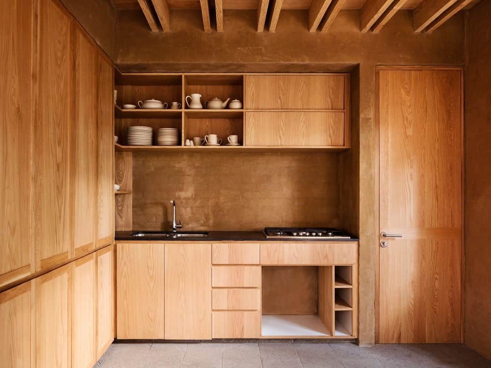 这是一个迷人的厨房，以木制橱柜为主，墙壁上布满了裸露的木制橱柜、抽屉和架子，反映了木质天花板和相同色调的木质裸露横梁。这呈现出一致的色调，与纹理石材地板很好地搭配。