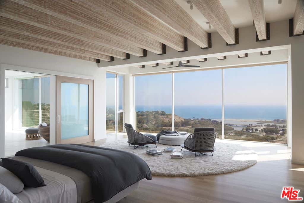 这间主卧室的亮点是美丽的海滨景色，大玻璃墙带来了丰富的自然光，照亮了天花板裸露的木梁的木质色调，硬木地板在休息区覆盖着毛茸茸的区域地毯。