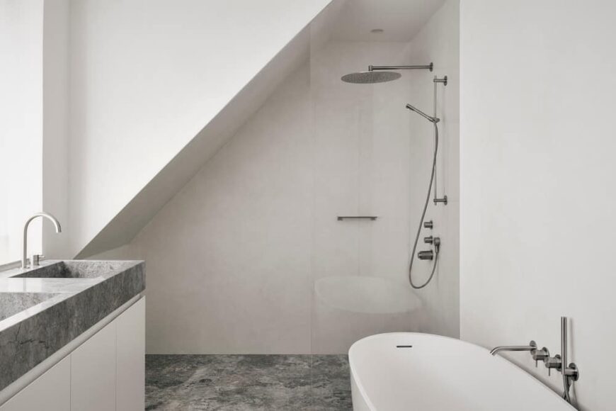 这是一间极简主义风格的主浴室，远侧有一个玻璃封闭的淋浴区，旁边是独立浴缸，对面是白色梳妆台，灰色台面与地板相配。