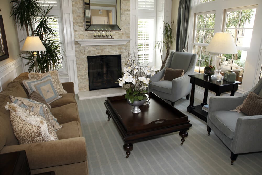 传统的客厅设计，华丽的暗木咖啡桌，两张蓝灰色扶手椅和一张棕色沙发。空间的两面墙都有窗户，为客厅提供自然光线。
