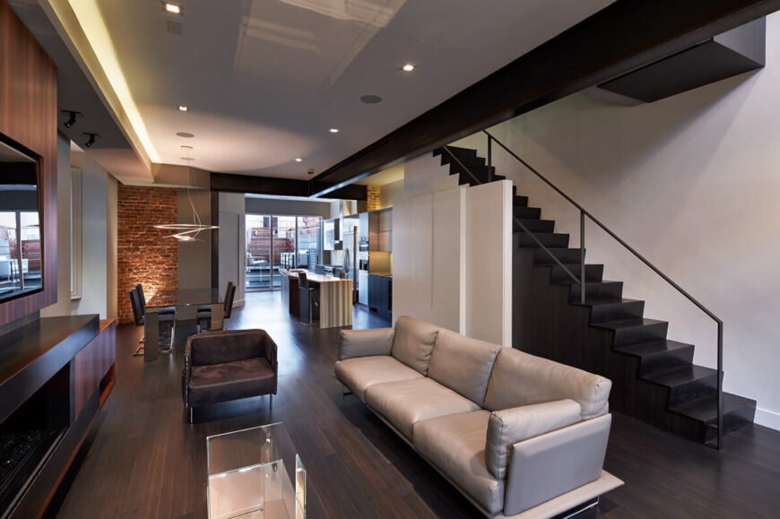 这是共享空间的相反角度，包括客厅、餐厅和厨房区域。黑色楼梯与天然硬木地板、白色墙壁和橱柜形成鲜明对比。浅色和深色皮革座椅环绕镜面玻璃咖啡桌。