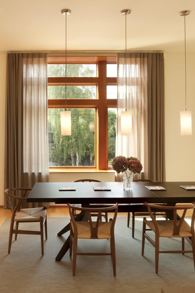 用餐区近景，深色木桌搭配浅色天然木椅，米色地毯。另一个天花板高的窗户提供自然光线，在悬挂的最小枝形吊灯后面。