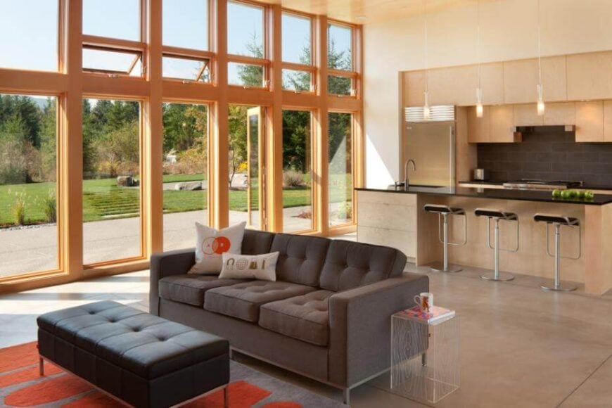 扩大的大开放空间与厨房和起居区共享，设有棕色沙发，玻璃木纹边桌，黑色皮革脚垫在灰色和橙色图案的地毯上。