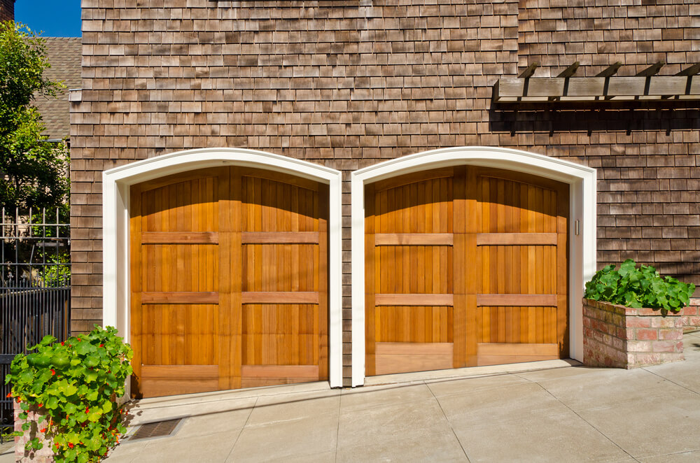 这个侧向的车库有两个浅色的天然木门，拱形，白色框架。定制形状适应倾斜车道。