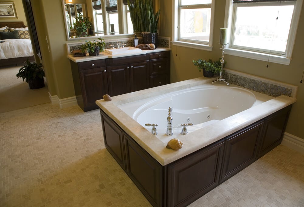 在浴室里，有米黄色的瓷砖地板和深色的木橱柜，这个椭圆形的浴缸配有漩涡喷嘴，坐落在凸起的表面，详细的瓷砖图案贴在外墙上。