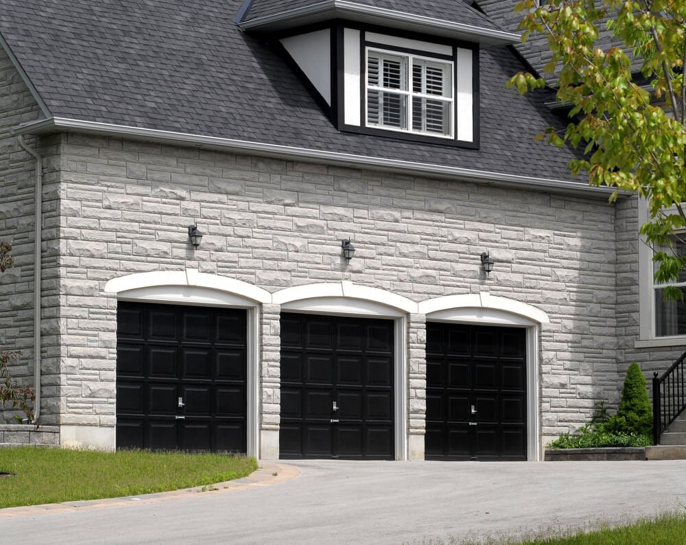 白色石砖住宅设有三个车库，拱形石门楣和金属灯烛台下的黑色门板。