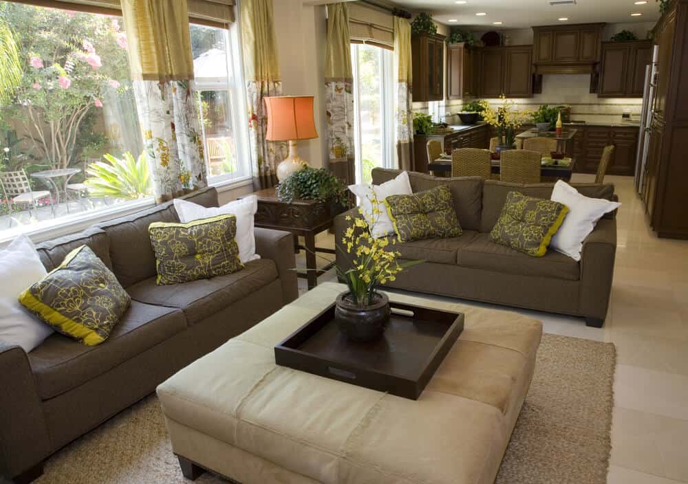 漫长的开放式客厅空间与远处的u形厨房共享，设有一对棕色沙发，面向大型方形米黄色皮革脚垫，带有可拆卸的深色木托盘桌。