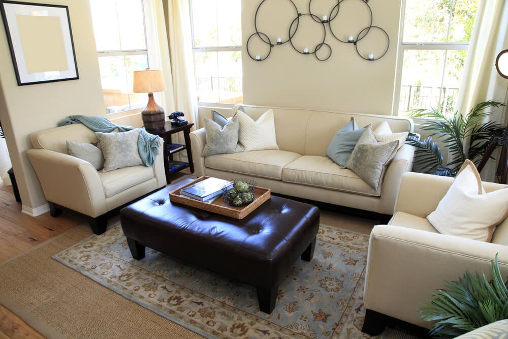 舒适的白色客厅，天然硬木地板和亚麻软垫座椅，围绕着大型长凳风格的纽扣簇绒棕色软凳。