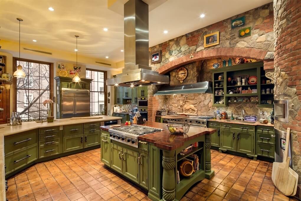 错综复杂的乡村厨房，砖和石头的工作。橱柜是有纹理的绿色。厨房布局杂乱，有几个工作区。