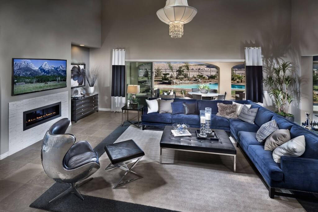 宽敞、开放的客厅铺着灰色瓷砖地板，配有蓝色织物组合沙发和铬背旋转扶手椅，中央有大型金属框架黑色簇绒皮革方凳。