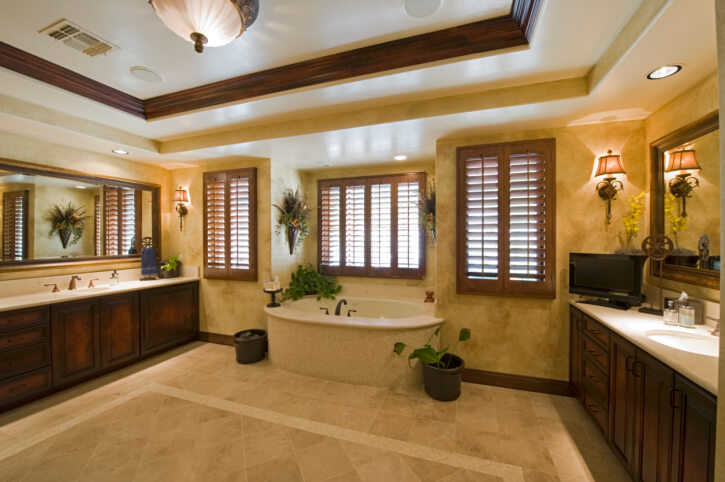 这间天花板很高的浴室铺着大面积的米黄色瓷砖地板，中间是与墙壁宽的深色木质梳妆台，白色大理石台面和中央的大型浴缸。