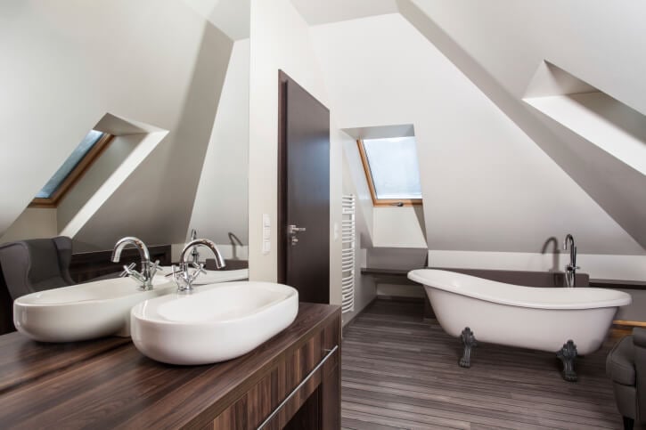 这是另一间浴室，大胆的白色倾斜天花板与深色天然木地板形成对比。梳妆台的色调较轻，有容器水槽，对面是传统的爪脚浴盆。