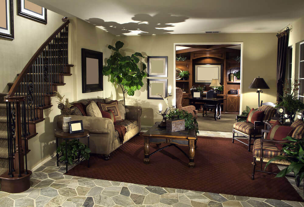 这间舒适的客厅位于左侧的雕花木和锻铁楼梯栏杆下，石瓷砖地板上铺着深紫红色地毯。