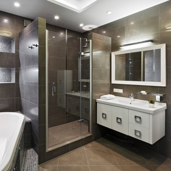 丰富的深棕色瓷砖地板和墙壁包裹着这个紧凑的浴室，在整个浴室中都有明亮的触感:角落浴缸，镜框和漂浮的木质梳妆台都是白色的。