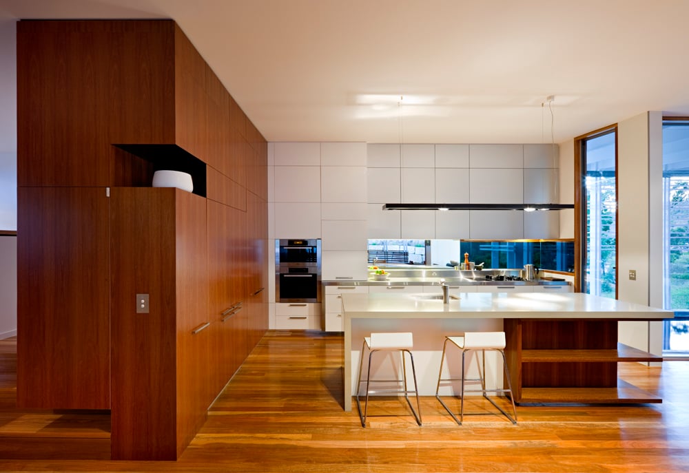 厨房充斥着天然木材和光滑的白色表面的对比，以带有内置搁架和用餐空间的大型白色和木质岛台为例。极简主义的橱柜设计与左侧的木镶板相辅相成。