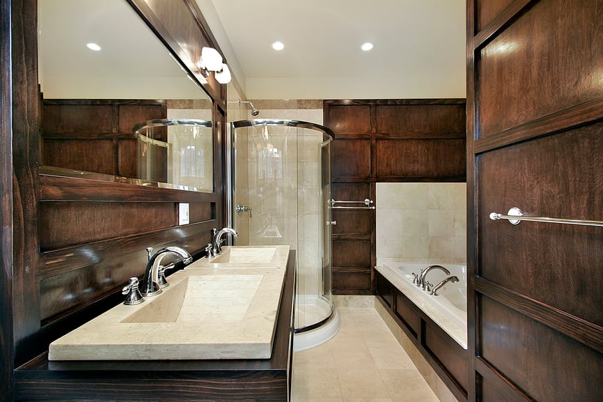 这个主浴室与墙壁和梳妆台上茂密的深色木镶板和白色大理石水槽、地板和浴缸形成对比。圆形玻璃淋浴架在角落里，有更多的米色瓷砖。
