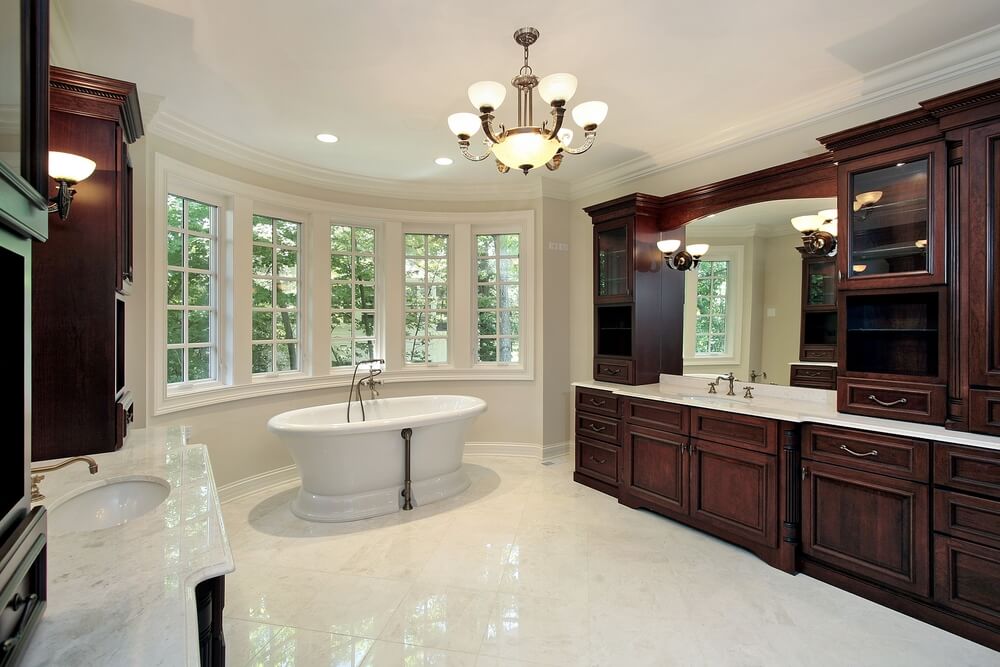 这间白色大理石浴室里有一对巨大的深色木质梳妆台，大理石台面上有一系列围绕镜子的橱柜。白色底座浴缸位于弧形窗户前的中央。