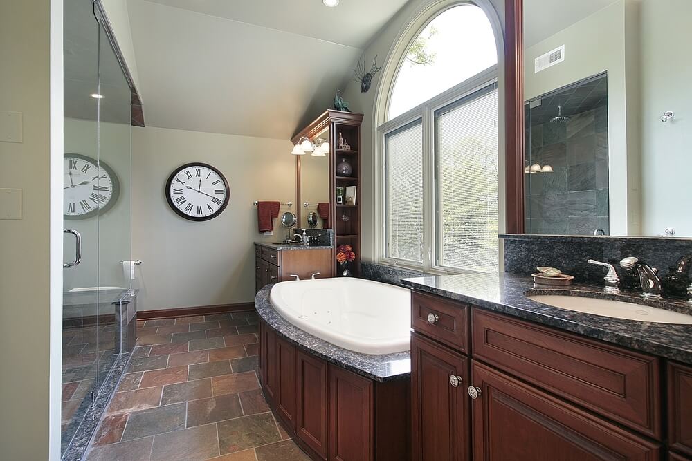 深色木质的双梳妆台位于配套的浴缸旁边，带有深色大理石台面。大的全玻璃淋浴架在黑色的石头瓷砖地板上。