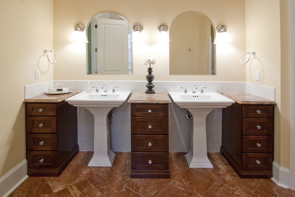 浅黄色色调的浴室覆盖着棕色大理石地板，在三个带大理石台面的深色木质梳妆台之间设有一对底座水槽。
