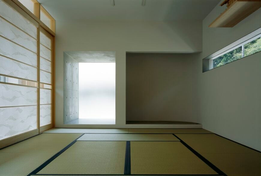 室内房间以席子地板、白墙和天然木材为特色，包括右侧狭缝窗上方的高架子。