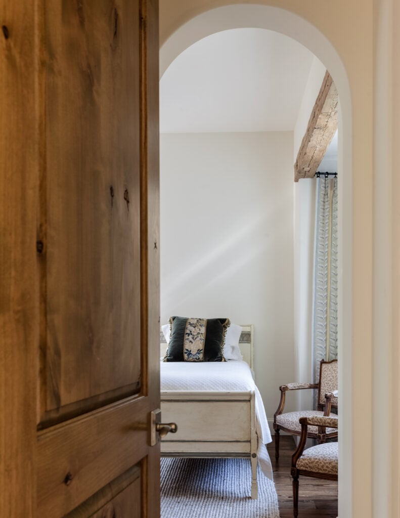 这间客卧，通过另一个拱形的门口，有更多的硬木地板，加上老式的木架扶手椅和一张乡村风格的床。