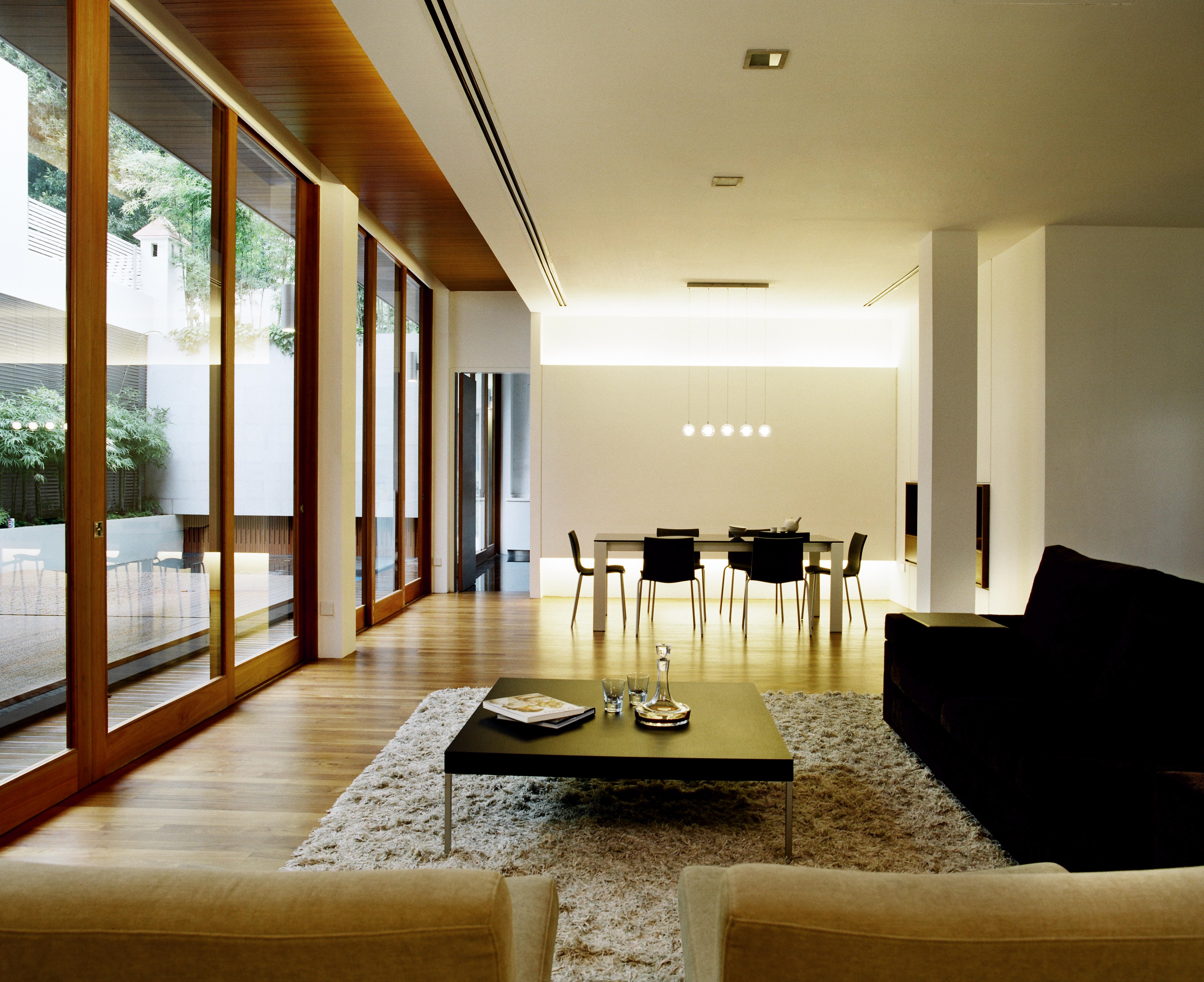 客厅位于与用餐区共享的开放空间，左侧有天然木框架玻璃露台入口。黑色沙发和一对驼色的椅子放在硬木地板上的厚地毯上，中间是金属和黑色木材的咖啡桌。