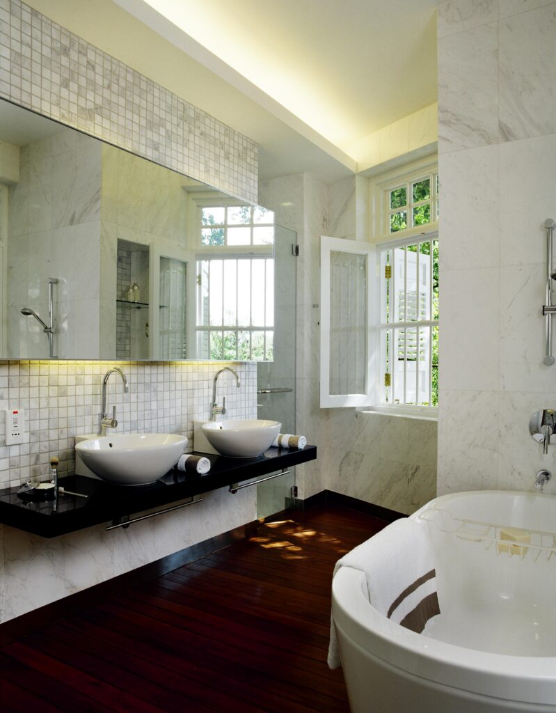 主浴室沐浴在白色大理石墙壁上，地板是深色的硬木地板。右边的底座浴缸对面是黑色的浮动梳妆台，带有一对容器水槽。玻璃门淋浴站在阴影白色框窗旁边。