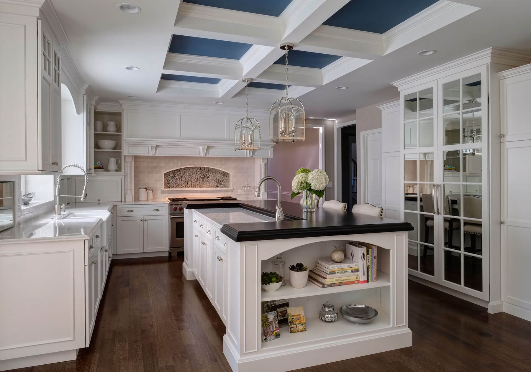 白色的厨房是一个巨大的黑色顶部的岛台，下面是蓝色镶板的天花板。镜面冰箱门横跨深色硬木地板，白色台面与岛台形成对比。