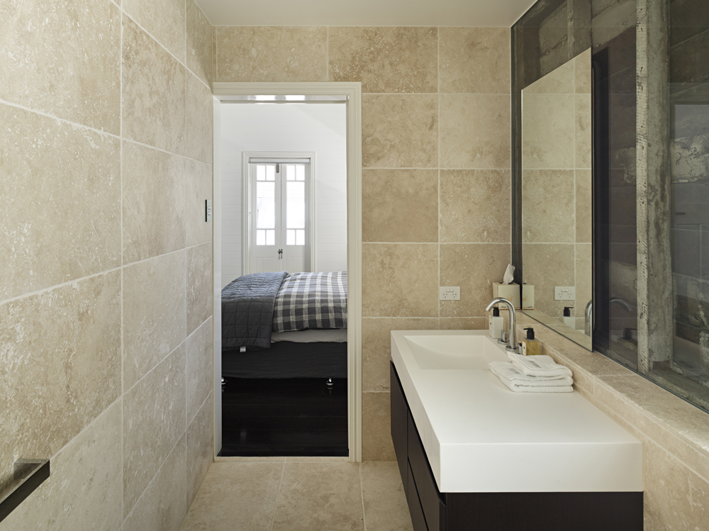 第二间浴室从地板到天花板都覆盖着米色瓷砖，以现代极简主义的深色木材梳妆台和白色台面为特色，右侧有滑动镜。