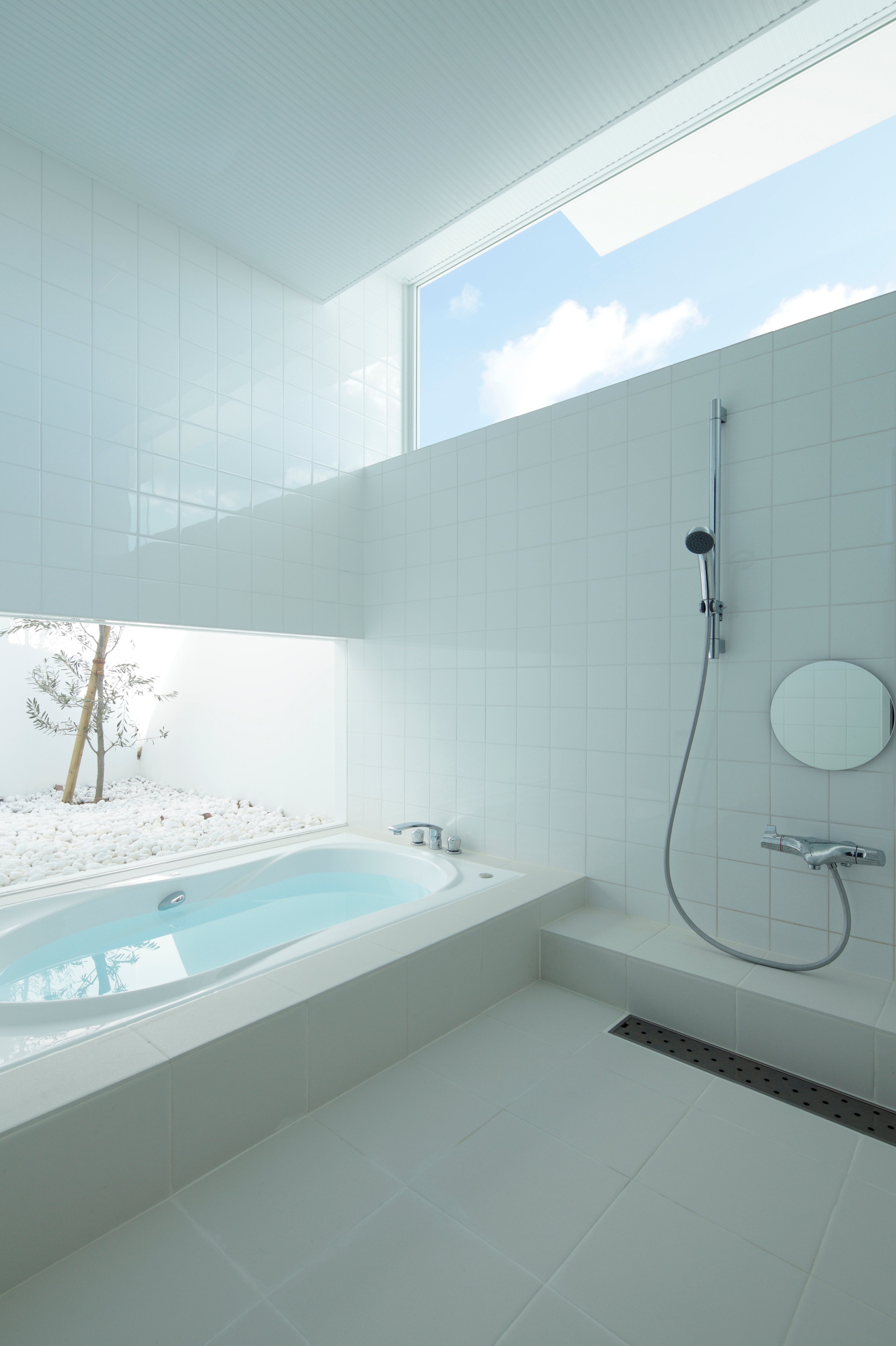 浴室是一个引人注目的、原始的白色瓷砖和阳光的广阔空间，开放式淋浴和按摩浴缸相邻，从左边较低的窗户可以看到外部的白色岩石花园。