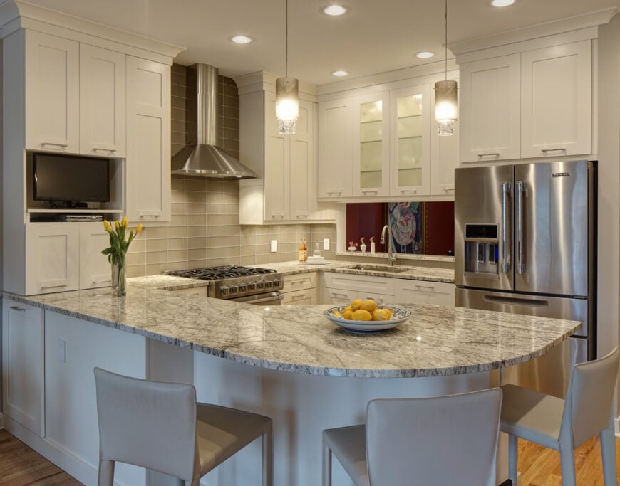 明亮的厨房设计以l形的台面包裹空间为特色，灰色花岗岩台面下方有弯曲的吧台式座椅。白色橱柜搭配钢制电器和米色瓷砖后挡板。