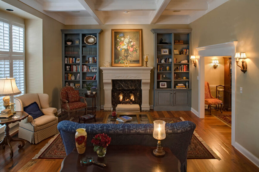 华丽的细节和多样的色调充斥着这个美丽的客厅，以米黄色墙壁上细致的白色天花板和丰富的硬木地板为特色。一对灰色的全高书架位于中央石壁炉的侧边，而蓝色、棕色和红色的椅子则分别围绕着带有镜子的咖啡桌的花纹地毯。