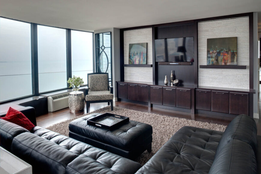 奢华的纽扣簇黑色皮革组合家具包裹着这个家庭活动室，炫耀硬木地板，深色木墙长架子，和棕色蓬松地毯。落地窗使空间更加明亮。