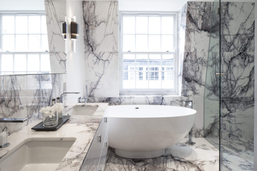 精致的纹理大理石填充了浴室，穿过玻璃淋浴，包裹着大窗户下的碗形底座浴缸。
