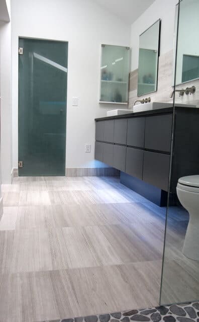 从淋浴门我们可以看到灰色的地板和木纹图案，黑色的梳妆台下面有LED照明和双容器水槽。