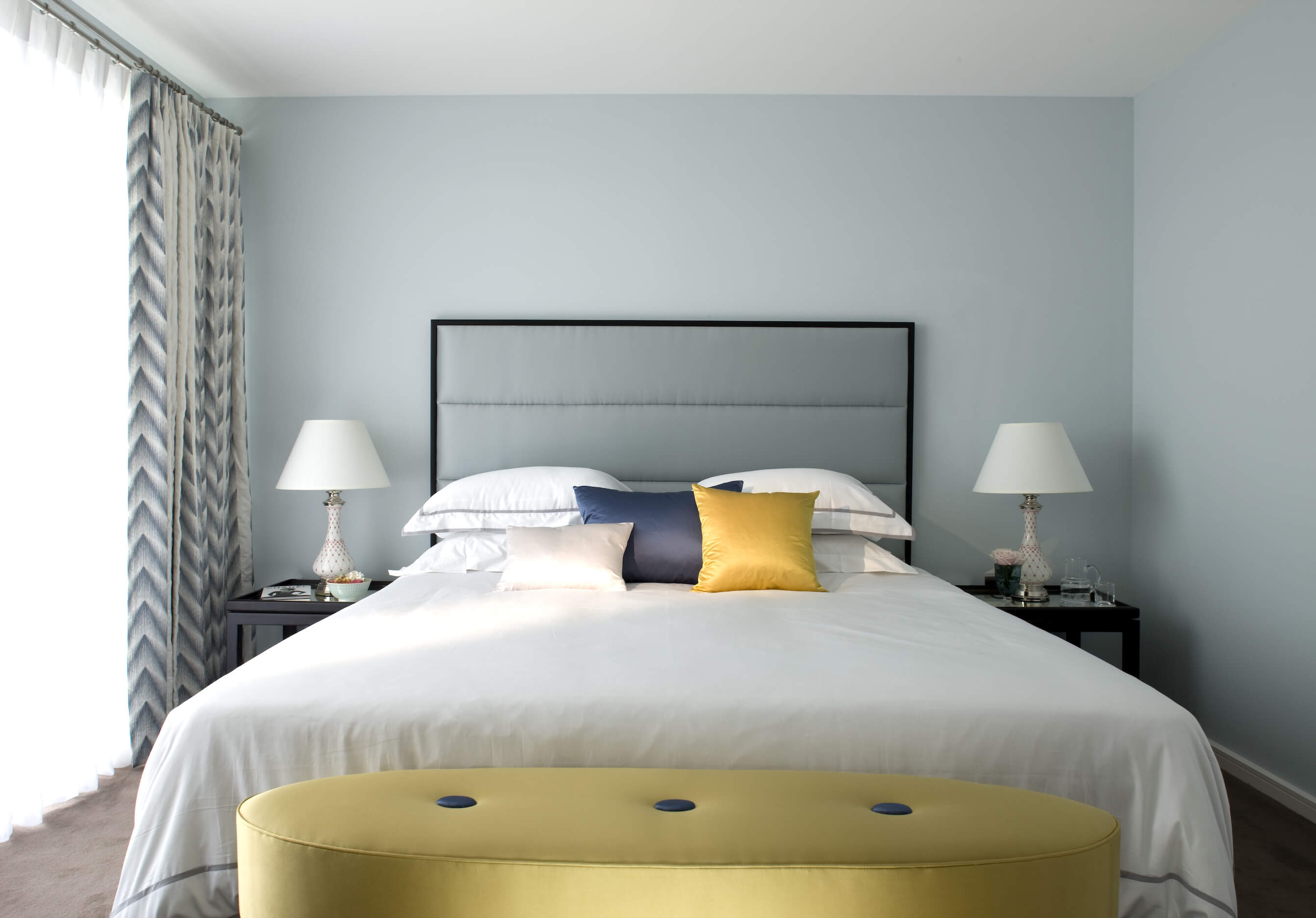 在卧室里，白色的金色色调与大胆的蓝色色调相结合，就像在床脚的枕头和椭圆形脚凳上看到的那样。柔和的天蓝色墙壁照亮了空间。