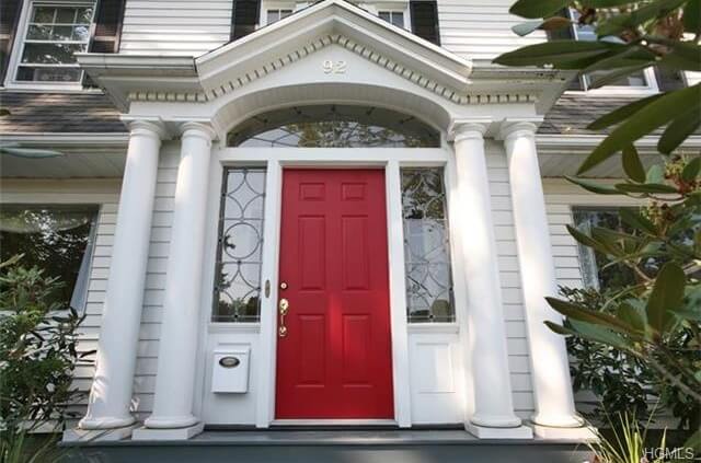这扇门是典型的村舍式前门。红色的前门有长方形的嵌板，金色的把手和锁。两侧的侧灯都有几何设计的透明玻璃板。横梁也是由透明玻璃制成，并连接到白色框架上。红白相间的小屋风格的门上有四根柱子，左边的侧灯下面有一个邮箱。