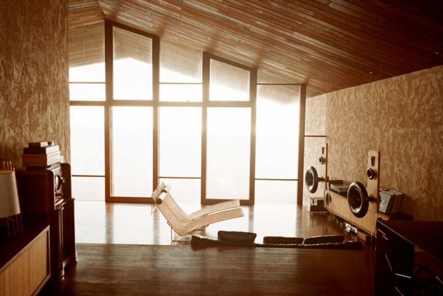 向下看客厅，我们看到一对躺椅面向定制的木质和外部扬声器音乐设置，阳光透过木质框架的全高玻璃照射。