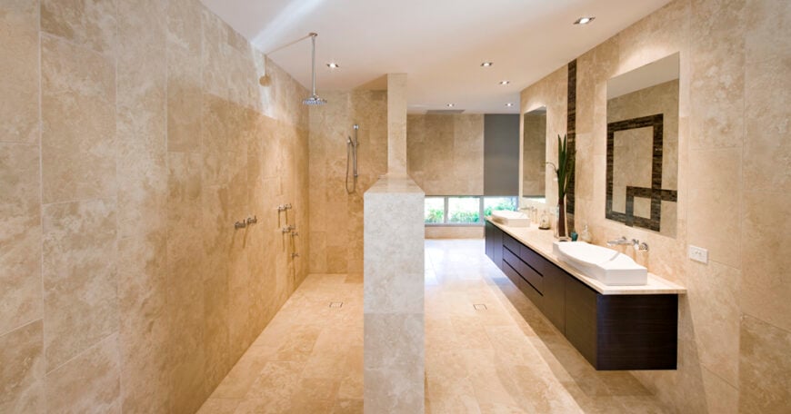 浴室从头到脚都铺着米黄色的大理石瓷砖。漂浮的深色木质双梳妆台和白色容器水槽悬挂在一对无框镜子下，而开放式设计的淋浴在左侧。