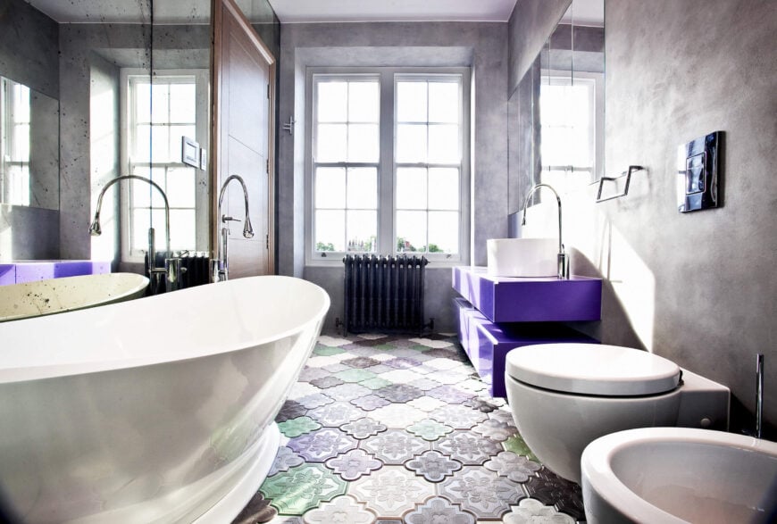 我们参观的第一间浴室以明亮的紫色梳妆台和五颜六色的纹理地砖为主。一个巨大的底座浴缸位于左侧的镜子下方，与柔和的墙壁和明亮的色彩细节形成对比。