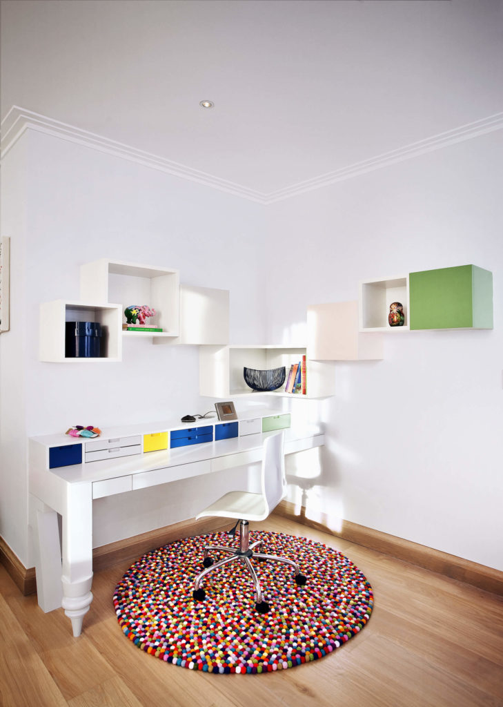 家庭办公空间里有一张极简主义的壁挂式白色办公桌和立方体架子，硬木地板上点缀着缤纷的色彩。明亮的、五彩缤纷的口香糖球地毯支撑着时髦的当代办公椅。