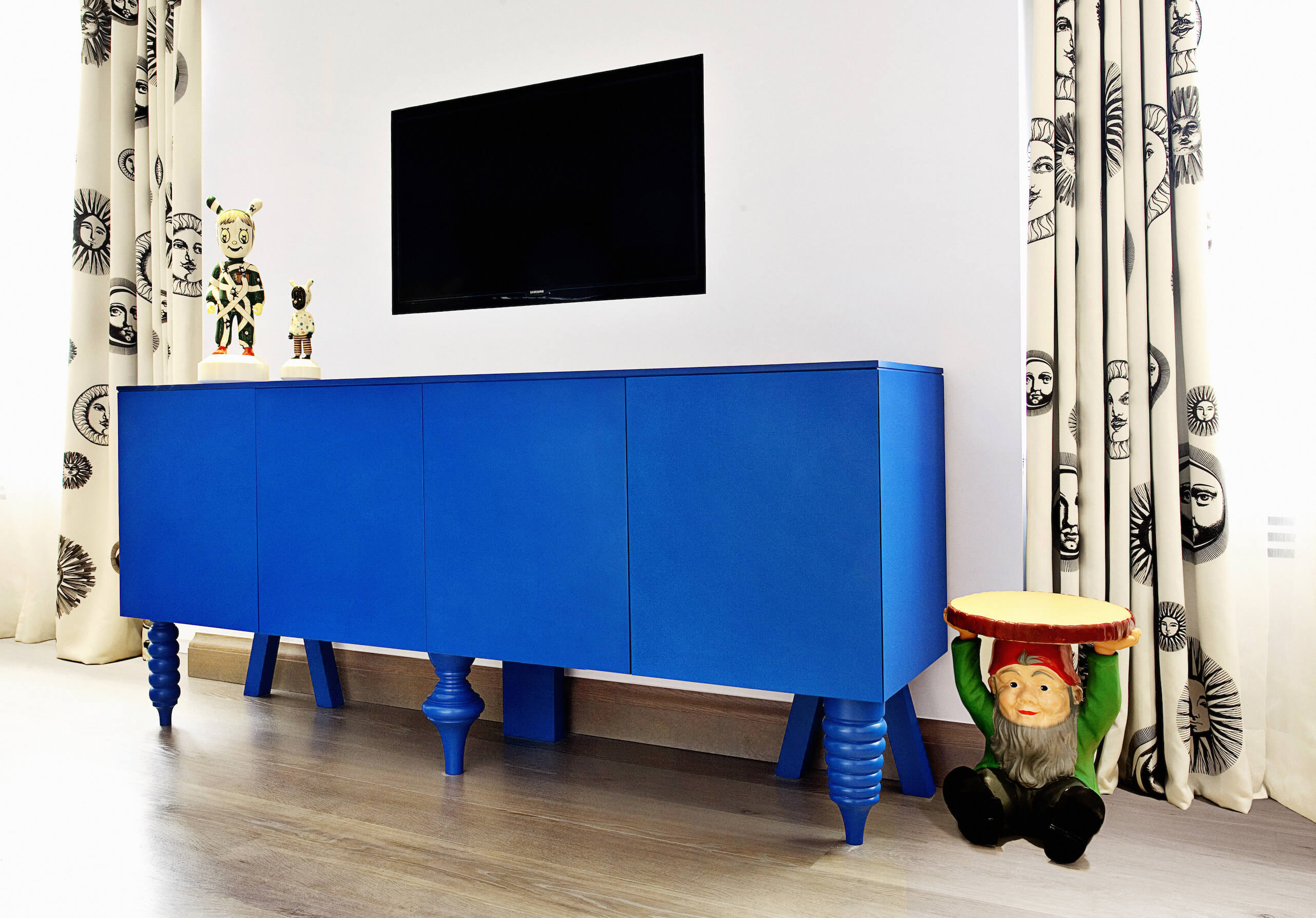 卧室里摆放着另一件大胆的家具;这个亮蓝色的橱柜站在阳光图案的窗帘之间，腿的设置不匹配。壁挂式电视悬挂在空间上方，展示着另一张侏儒桌子和一对小雕像。