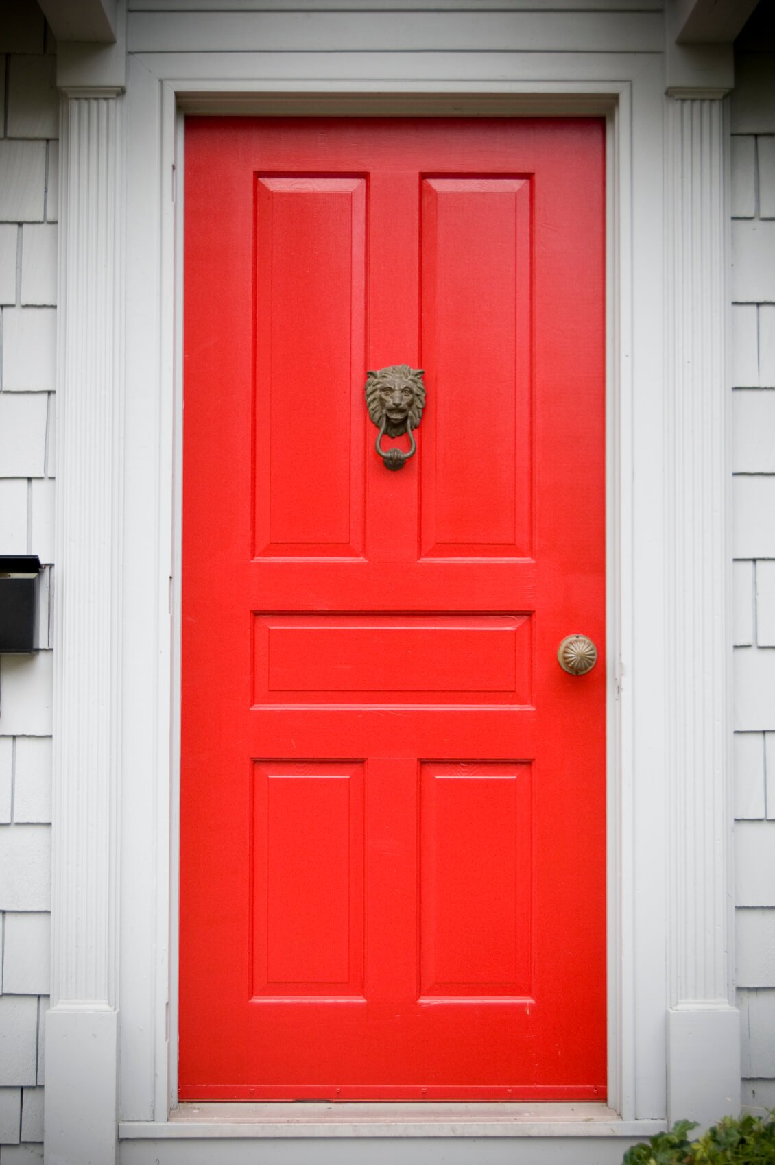 米白色的造型和框架支撑着这扇亮红色的门，它有五个矩形面板，并有一个青铜门环和一个青铜门把手。门的左边有一个固定在墙上的黑色邮箱。