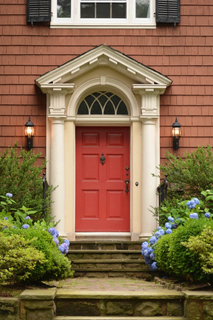 通往大胆的红色前门的石头走道和台阶两旁种着灌木和亮紫色的绣球花。房子的壁板是暗淡的砖红色。