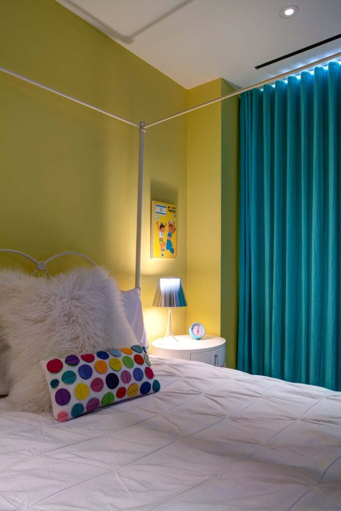 这是孩子们的一间卧室，墙壁是黄色的，窗帘是水绿色的，床上用品和床头柜都是白色的。这里出现了彩虹色的圆点图案。