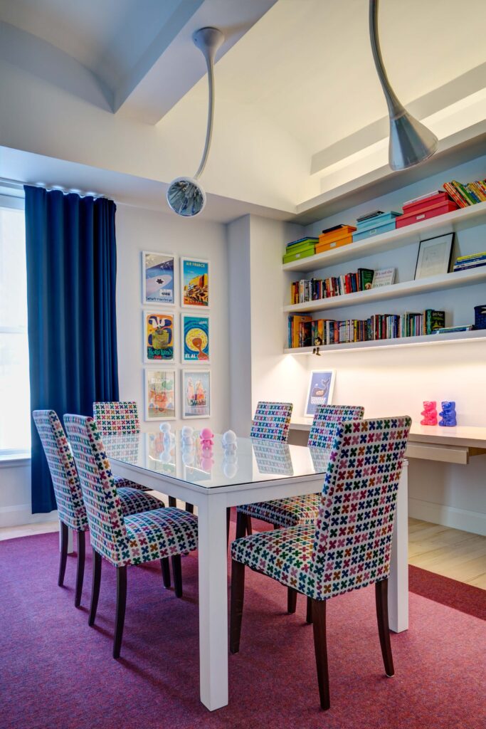 宽敞的游戏室拥有明亮和色彩的完美结合，白色的玻璃桌面桌子周围环绕着彩虹图案的强调椅。内置的书架上放着书，旁边是墙上的艺术品。