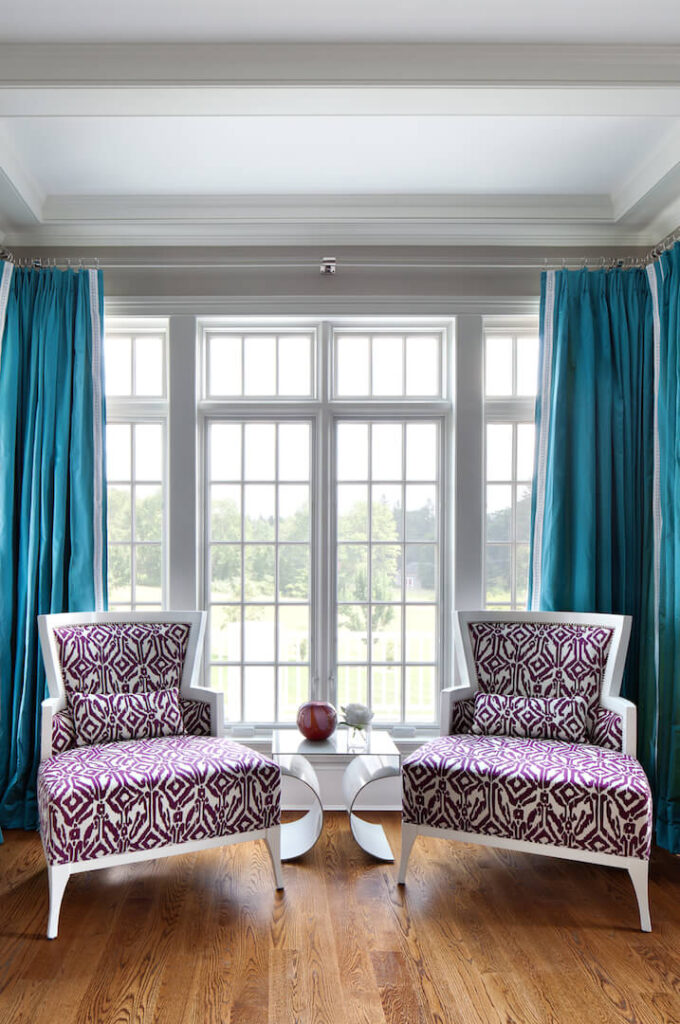 仔细看白色和紫色的客厅在大窗户前。两把椅子之间是一张现代的边桌，上面有几件小装饰品。椅子后面大胆的绿松石色窗帘是一个大胆的颜色宣言。